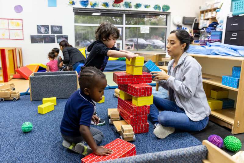 A classroom scene featuring a woman sitting on the floor, assisting a young boy as he builds a tall tower with colorful blocks. Nearby, another young child is playing with smaller blocks. In the background, additional children are engaged in various activities, and the room is filled with educational toys and materials. The atmosphere is lively and playful, reflecting a nurturing learning environment.