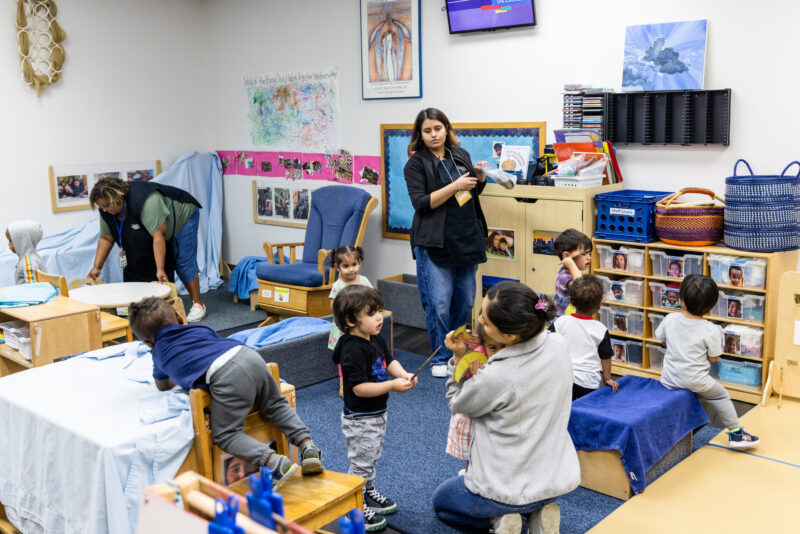 Alt text: A colorful classroom filled with young children engaged in various activities. Some children are playing on small wooden chairs and tables, while others are interacting with caregivers. One caregiver is reading to a group of children, and another is assisting a child. The room is decorated with educational posters and has shelves filled with toys and books.