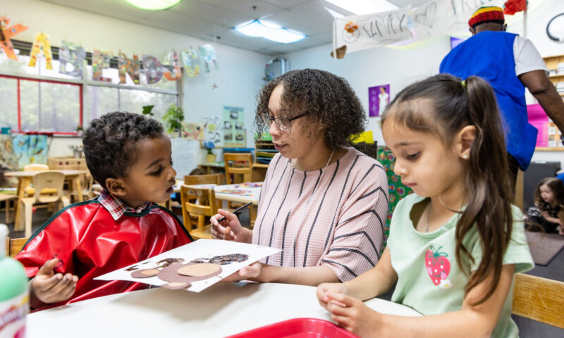 A teacher sits at a table with two young children in a colorful classroom. One child, wearing a red smock, is showing a craft project that includes various shapes, while the teacher attentively engages with him. The other child, wearing a light green shirt with a strawberry design, watches with interest. In the background, another adult is seen assisting other children in the classroom, which is decorated with artwork and educational materials.