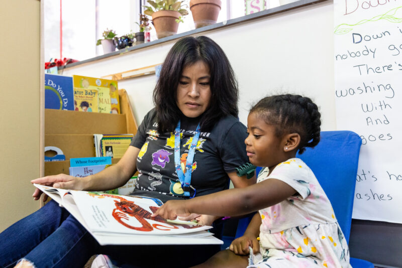A woman and a young girl are sitting together in a cozy reading area. The woman, wearing a colorful t-shirt, is engaged in reading a large picture book with the girl, who is pointing at an illustration. The background features shelves with books and potted plants, creating a warm and inviting atmosphere for reading.