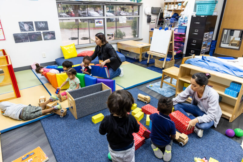 Alt text: A colorful early childhood classroom filled with children engaged in play. Several toddlers are playing with building blocks and toys on a soft blue carpet. Two caregivers are interacting with the children, one assisting a child with a toy truck and another helping a group of kids with a colorful play structure. The room is bright and organized, with various educational materials and furniture in the background.