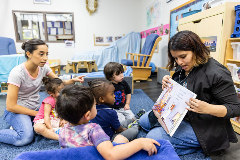 A caregiver is reading a storybook to a small group of young children sitting on a blue carpet in a classroom setting. One child is sitting on a cushion, while others are gathered around, attentively listening. Another adult is pointing towards the book, engaging with the children. The room is bright and decorated with educational materials and colorful artwork.