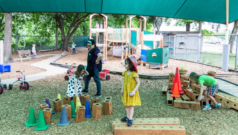 A vibrant outdoor playground scene under a large green canopy. Children are engaged in various activities: two girls, one in a yellow dress and the other in a polka dot outfit, are playing with wooden blocks and cones on the ground. An adult, possibly a caregiver, is supervising nearby. In the background, there is a wooden play structure with slides and climbing areas, and other children are playing on tricycles and exploring the space. The ground is covered with wood chips, creating a natural play environment.