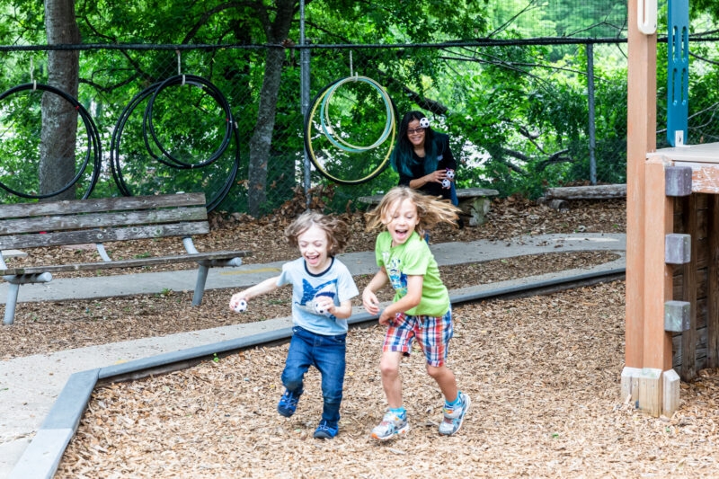 Two young children are running joyfully in a playground, with one child wearing a light blue shirt and the other in a green shirt. They have big smiles on their faces and their hair is flying as they play. In the background, a woman is sitting on a bench, watching them with a smile. There are hula hoops hanging on a fence nearby, and the ground is covered with wood chips.