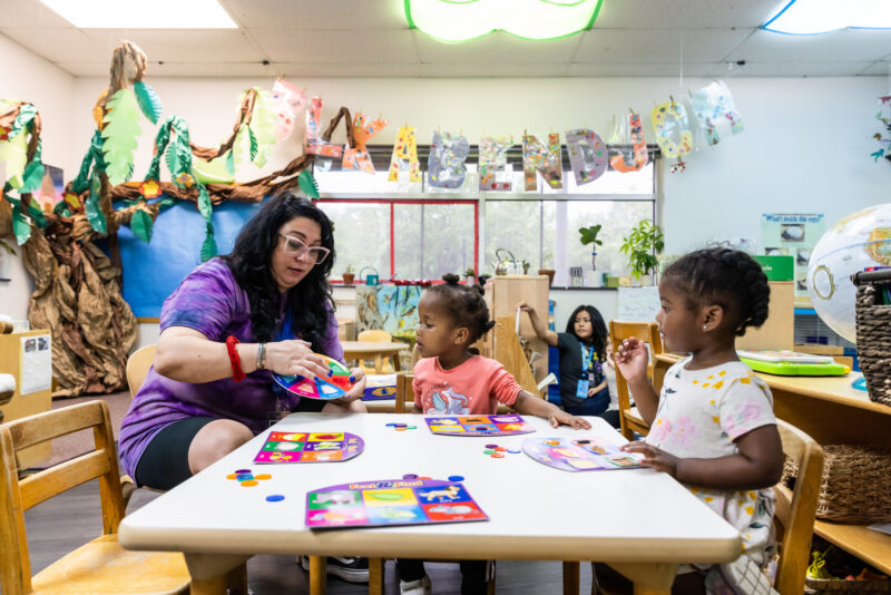 A classroom scene featuring a teacher interacting with two young children at a table. The teacher, wearing glasses and a colorful shirt, is showing a game or activity to the children, who are focused on the materials in front of them. One child is seated on the left, while the other is on the right, both engaged in the activity. In the background, another child is sitting quietly, and colorful decorations, including a banner with letters, adorn the walls. The atmosphere is lively and educational.