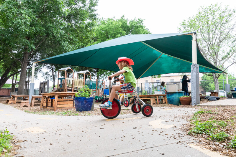 A young child wearing a red cowboy hat rides a tricycle along a paved path in a playground. The area is shaded by a large green canopy, and in the background, there are wooden play structures and tables surrounded by potted plants. Trees provide additional greenery, creating a lively outdoor environment.