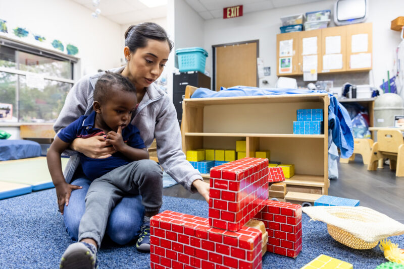 A caregiver is sitting on a blue carpet in a classroom, holding a young child on her lap. She is focused on building a structure with colorful blocks, which are red and blue. The classroom is bright and organized, with shelves and educational materials visible in the background. The child appears engaged and curious about the building activity.